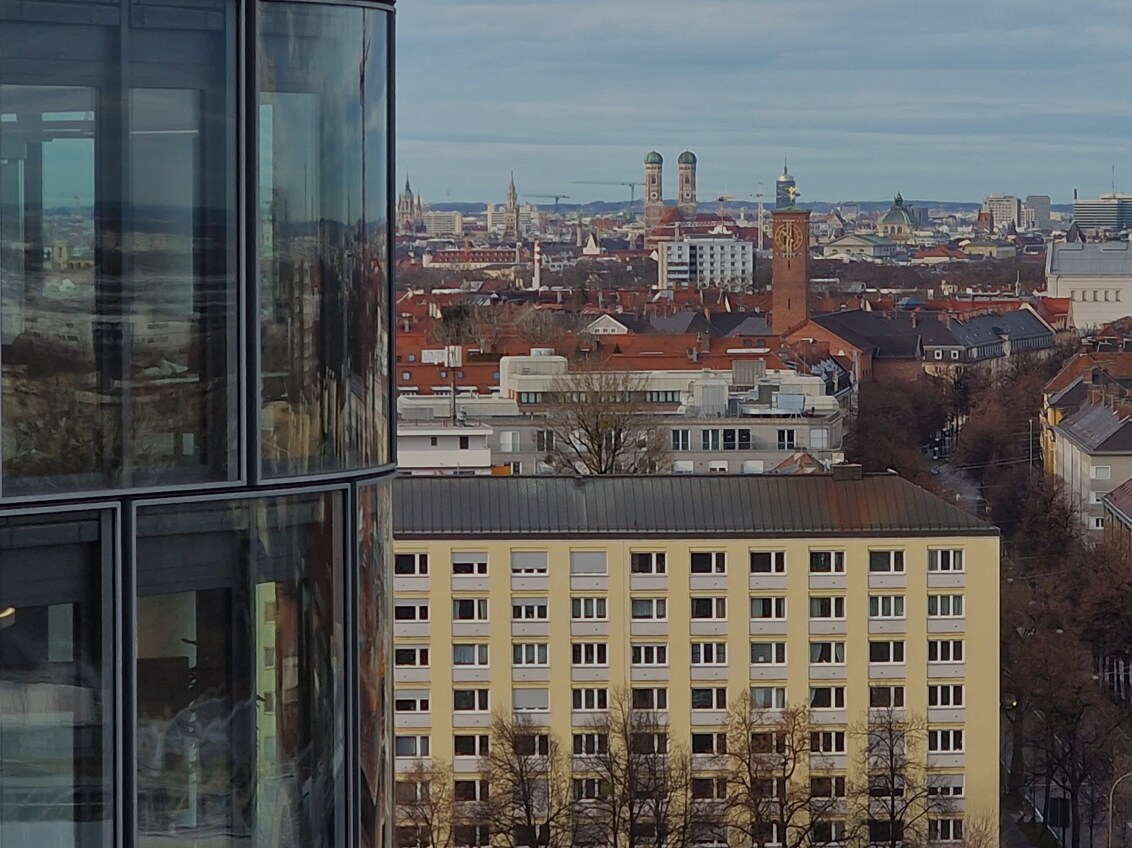 A scenic urban panorama of Munich featuring historic landmarks, red-roofed buildings, and a curved glass skyscraper in the foreground.