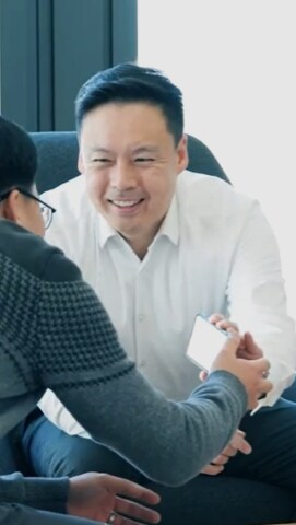 Two colleagues having a friendly conversation, with one showing something on a smartphone to a smiling man in a white shirt.