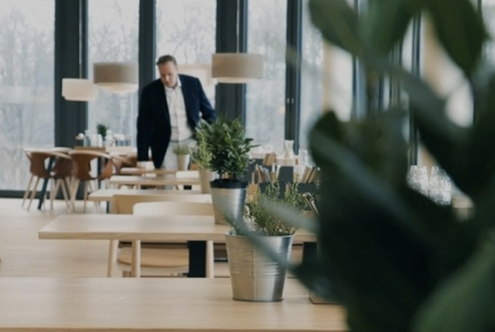 A modern, sunlit office cafeteria with minimalist wooden furniture and potted plants, as a man in a suit walks in the background.