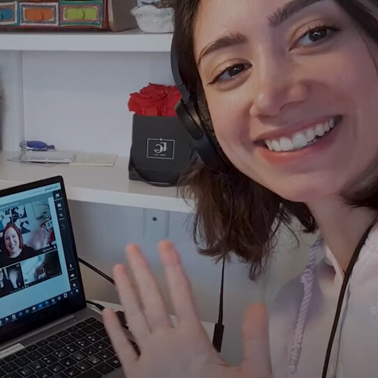 Smiling young woman wearing headphones waves at her laptop screen during a video conference call in a home office setting.