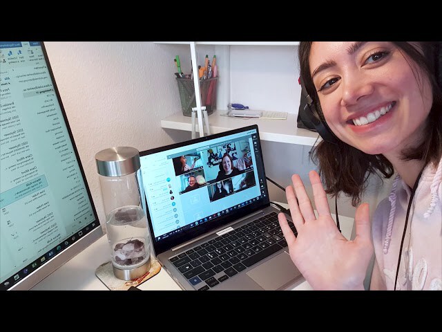 Smiling young woman wearing headphones waves at her laptop screen during a video conference call in a home office setting.
