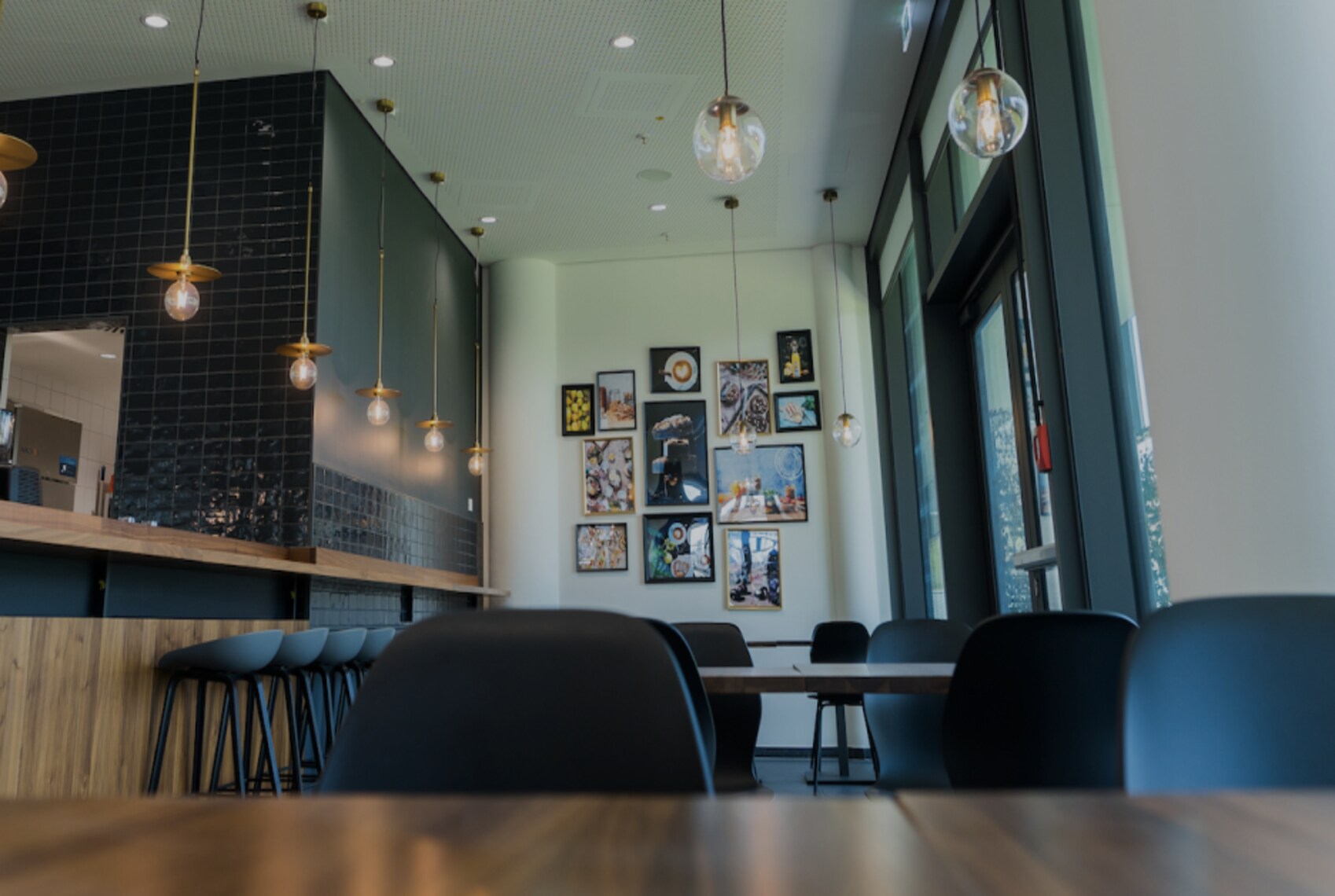 A stylish, modern café interior featuring pendant lights, black tile walls, high stools, and art-covered walls in a cozy, well-lit space.
