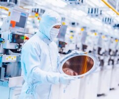 A technician in cleanroom attire examining a silicon wafer in a semiconductor foundry.