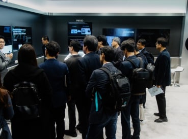 A group of conference attendees gathered around a speaker, listening to a presentation in a modern exhibit area with digital displays.
