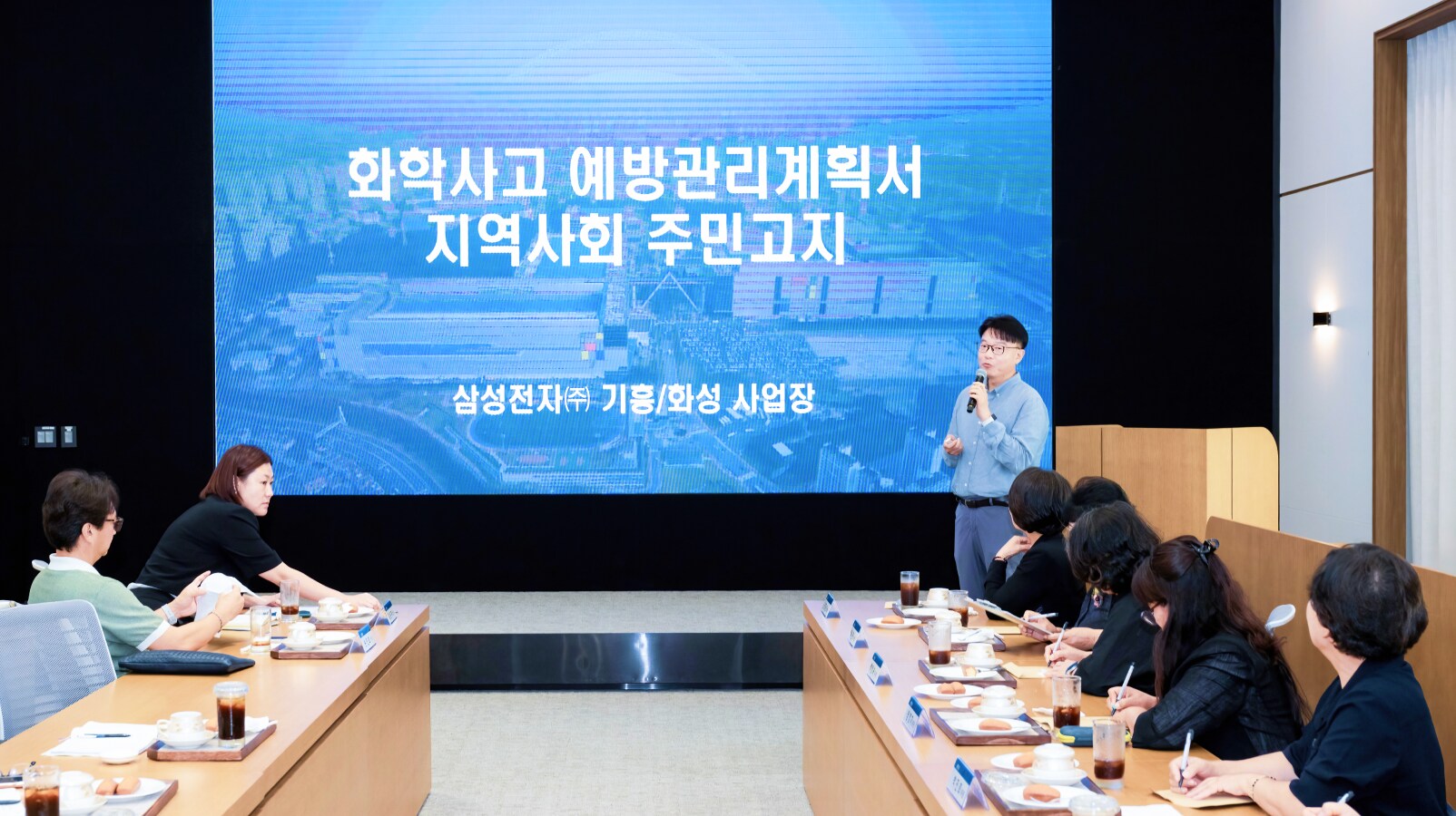 삼성전자 평택캠퍼스 회의실에서 열린 화학사고 예방관리계획서 지역사회 주민 고지 발표, 대형 스크린 앞에서 발표자가 마이크를 들고 설명하고 위원들이 긴 회의 테이블 양쪽에 앉아 경청하는 모습