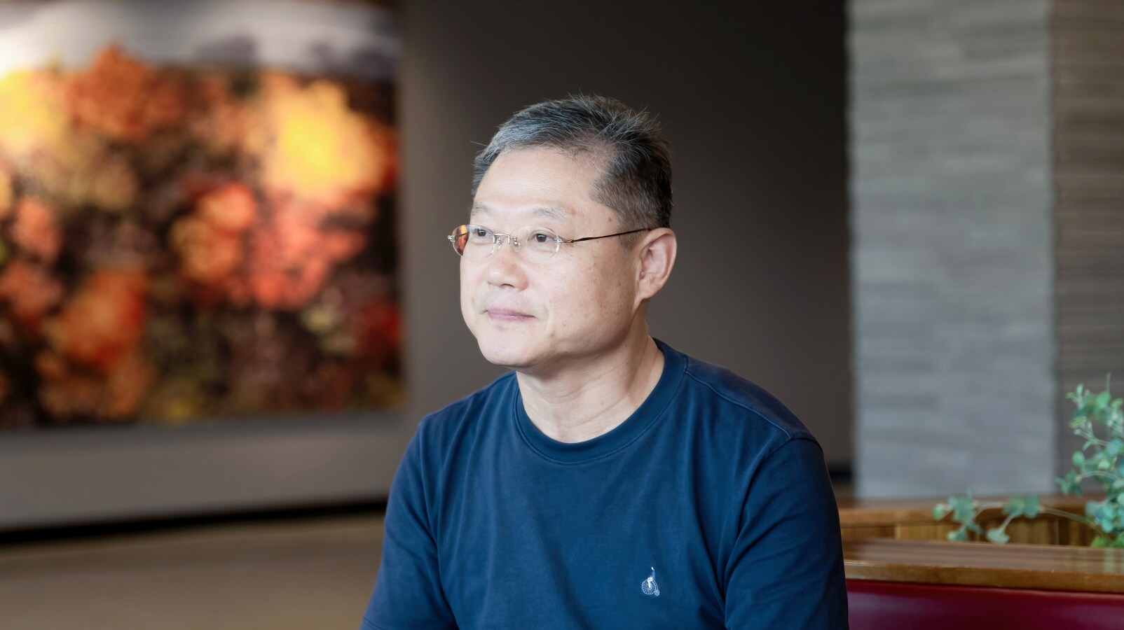 파란 티셔츠를 입은 김영길 동탄2동 주민자치회장이 실내 전시 공간에서 인터뷰 중 정면을 바라보며 미소 짓고 있는 모습. 뒤편에는 흐릿하게 보이는 대형 회화 작품이 걸려 있다.