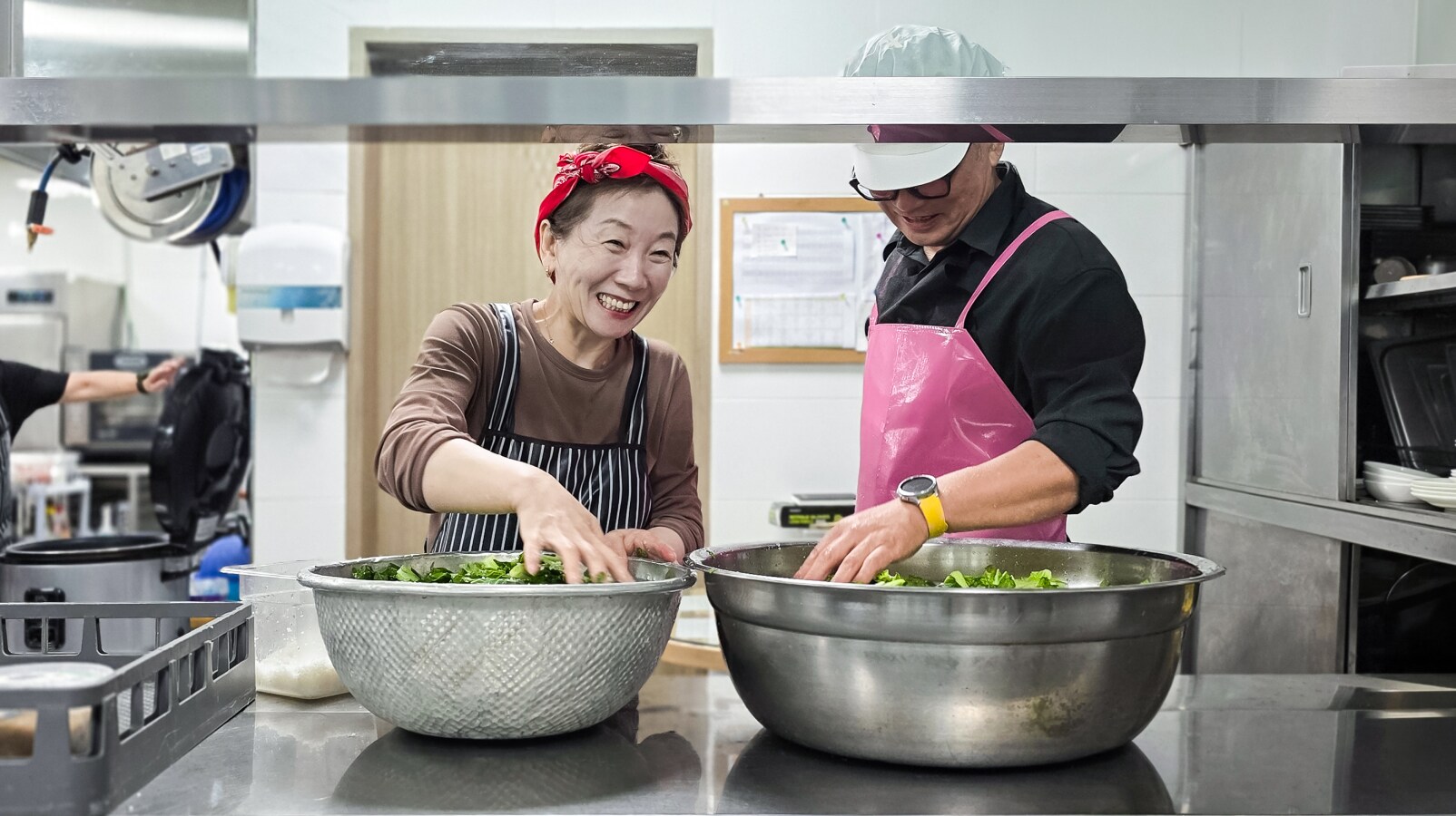 빨간색 머리띠를 하고 미소 짓고 있는 여성과 분홍색 앞치마와 위생모를 착용한 남성이 주방에서 커다란 금속 그릇에 담긴 채소를 소금에 절이고 있습니다.