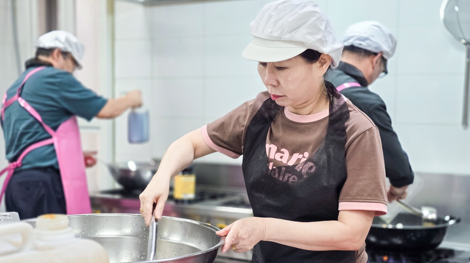 흰 모자와 검은 앞치마를 착용한 여성이 주방에서 커다란 금속 그릇 속의 내용물을 젓고 있으며, 배경에는 동료 두 명이 일하고 있습니다.