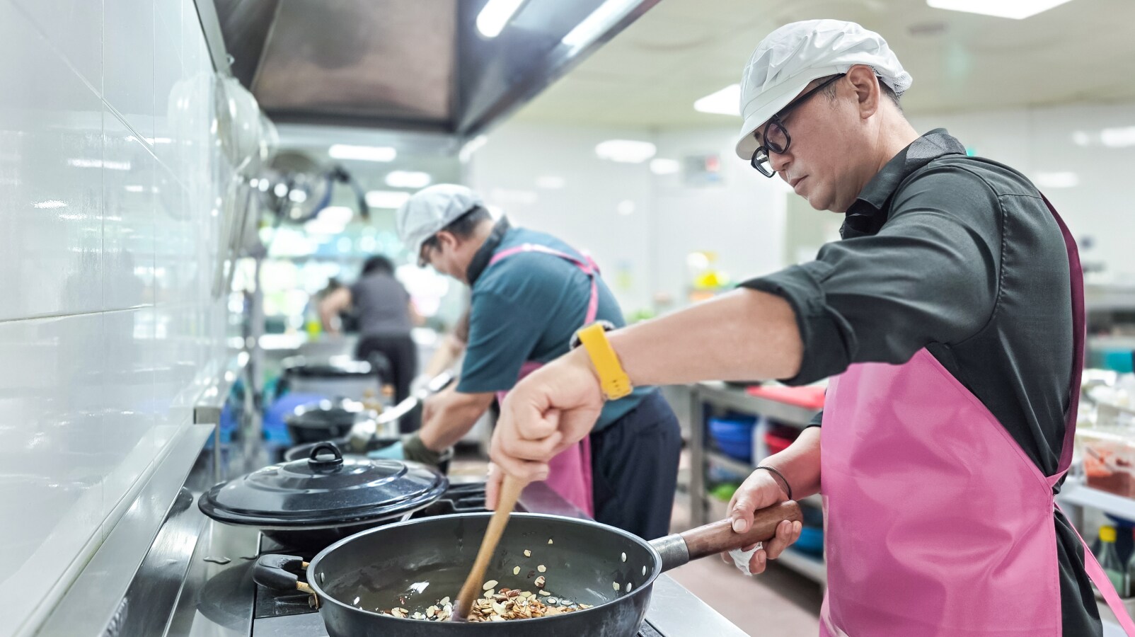 분홍색 앞치마를 두른 남성이 커다란 팬에 든 음식을 젓고 있고, 배경에서는 다른 동료들이 음식을 준비하고 있습니다.