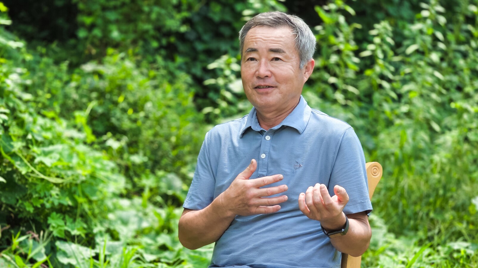 푸른색 폴로 셔츠를 입은 남성이 무성한 초록빛 식물을 배경으로 야외에 앉아 손짓을 섞어가며 이야기하고 있습니다.