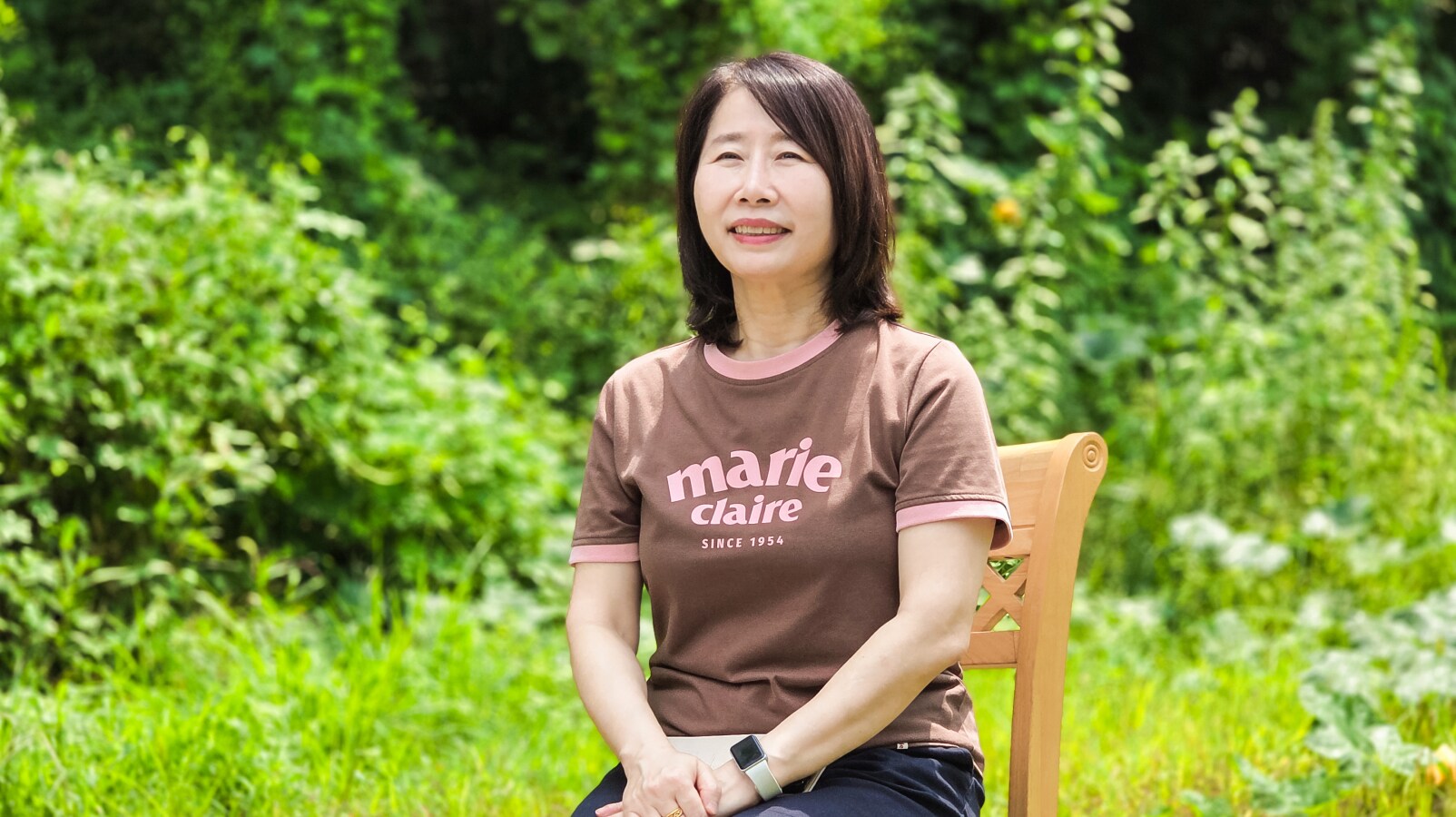 갈색 '마리끌레르(Marie Claire)' 티셔츠를 입은 여성이 무성한 초록빛 식물에 둘러싸인 야외의 나무 의자에 앉아 미소 짓고 있습니다.