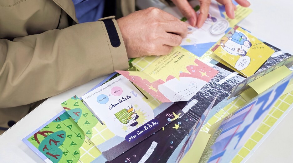 팝업북 페이지 위에서 손으로 캐릭터와 배경 종이를 조립하며 제작 체험을 진행하는 손동작 클로즈업