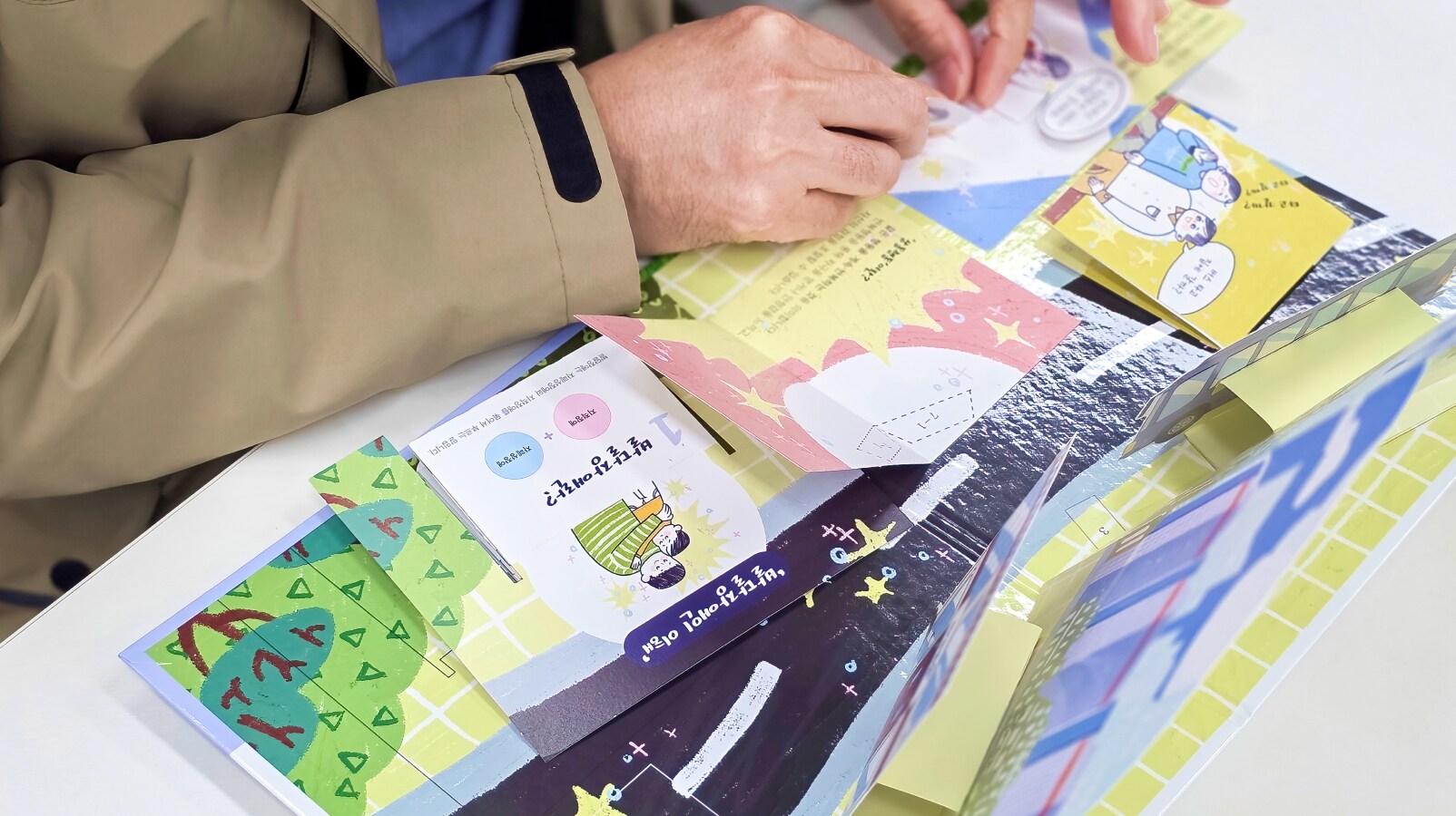 팝업북 페이지 위에서 손으로 캐릭터와 배경 종이를 조립하며 제작 체험을 진행하는 손동작 클로즈업