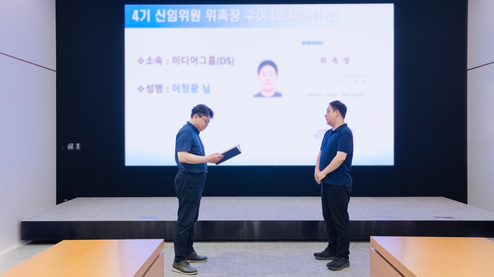무대 위 대형 스크린에 위촉장 이미지가 띄워진 가운데 마주 선 두 남성 중 한 명이 위촉장을 낭독하는 장면