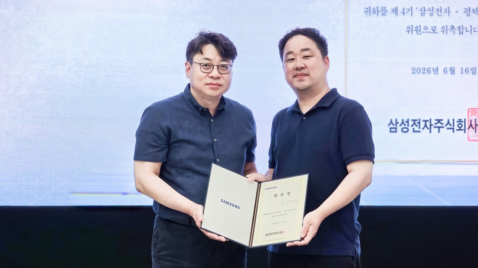 위촉장을 함께 들고 카메라를 향해 서 있는 두 남성의 기념촬영