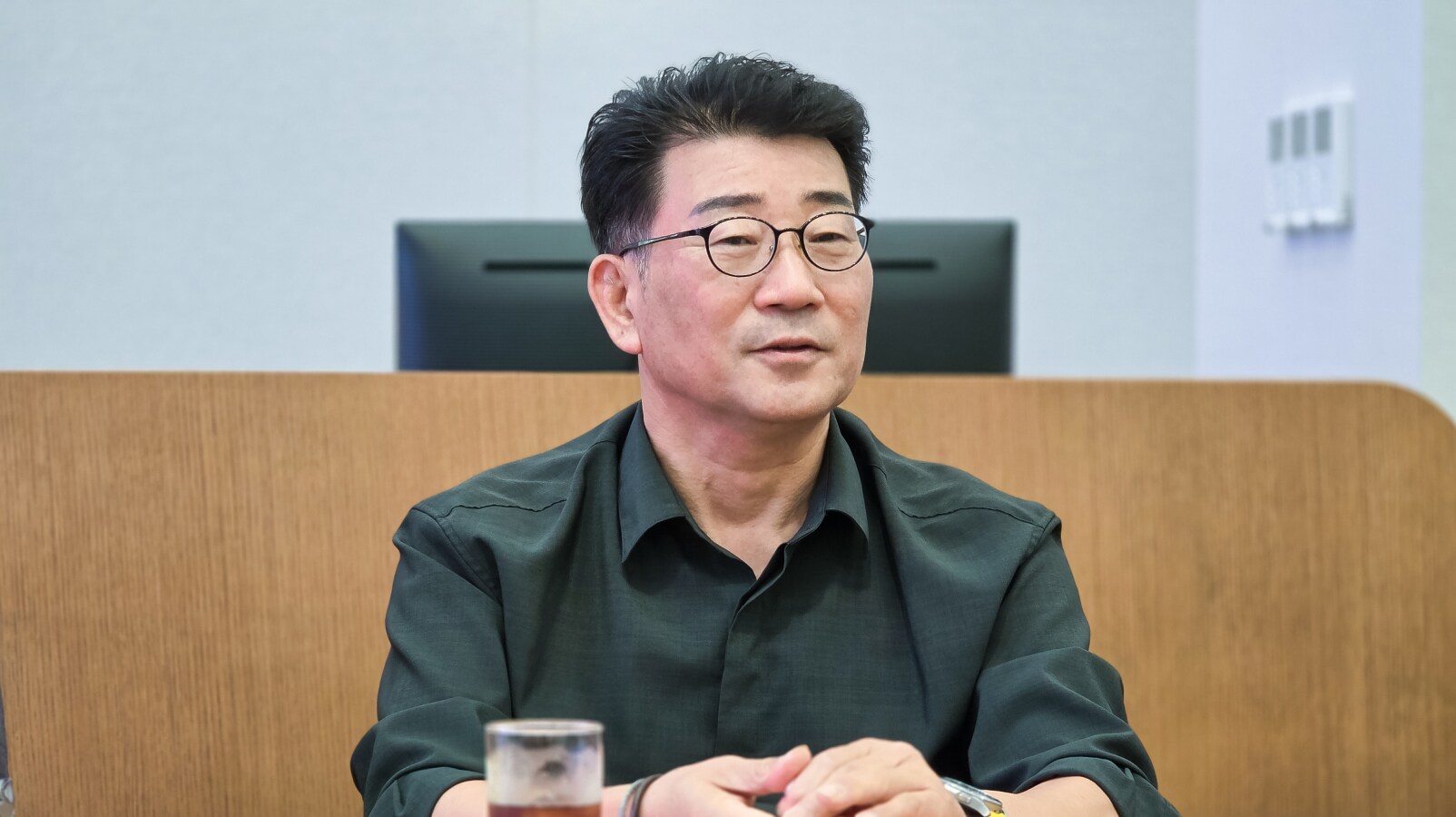 회의실 스크린 앞자리에서 안경을 쓰고 손을 모은 채 발언하는 남성