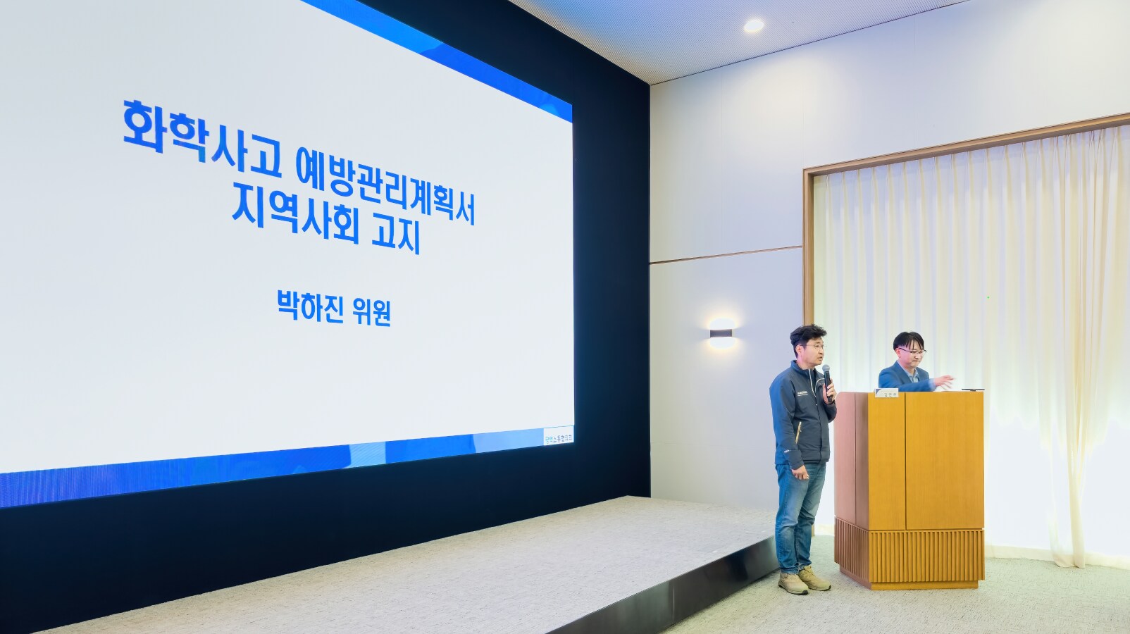 화학사고 예방관리계획서 지역사회 고지 제목이 띄워진 스크린 앞에서 마이크를 들고 발표하는 남성과 사회를 보는 또 다른 남성