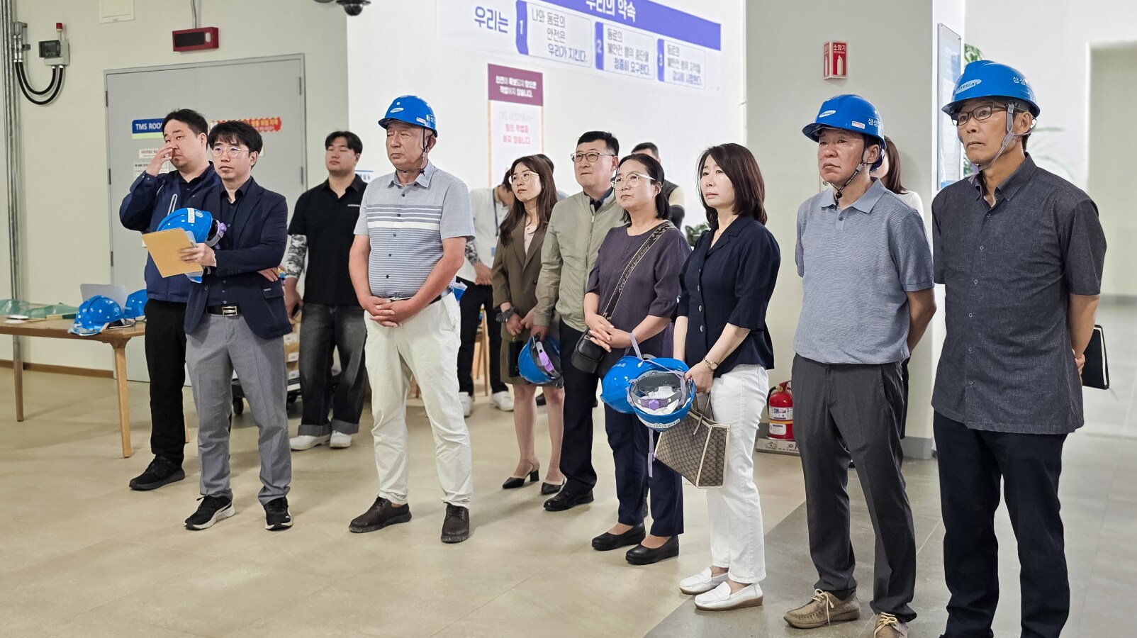파란색 안전모를 착용하고 벽면 안전 수칙 게시물 앞에 줄지어 서서 시설 설명을 듣는 사람들