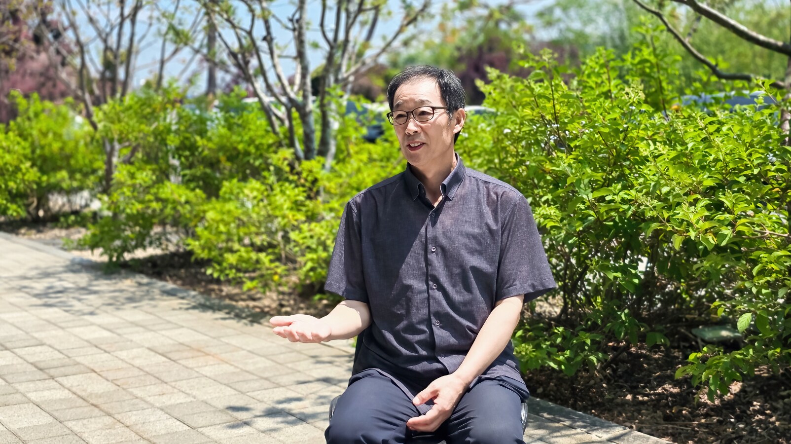 초록 나무가 우거진 야외 벤치에 앉아 손짓하며 인터뷰에 답하는 안경 쓴 남성