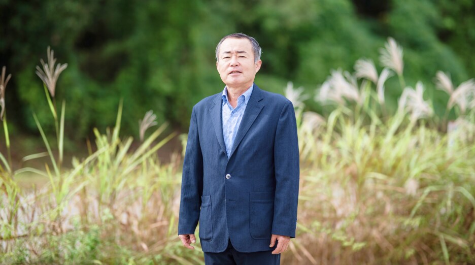 김재환 위원이 푸른 갈대밭 앞에서 정면을 바라보며 서 있는 모습. 이장협의회 사무국장으로서 주민들의 의견을 성실히 전달한 인물의 차분한 표정이 돋보인다.