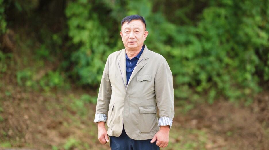 푸른 나무가 배경으로 보이는 야외에서 우제욱 대표위원이 정면을 바라보며 서 있는 모습. 밝은 표정으로 카메라를 향해 자연스럽게 포즈를 취하고 있다.