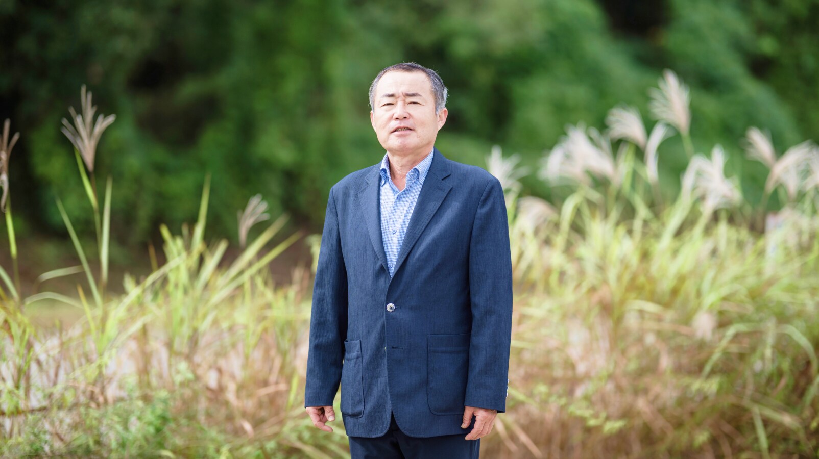 김재환 위원이 푸른 갈대밭 앞에서 정면을 바라보며 서 있는 모습. 이장협의회 사무국장으로서 주민들의 의견을 성실히 전달한 인물의 차분한 표정이 돋보인다.