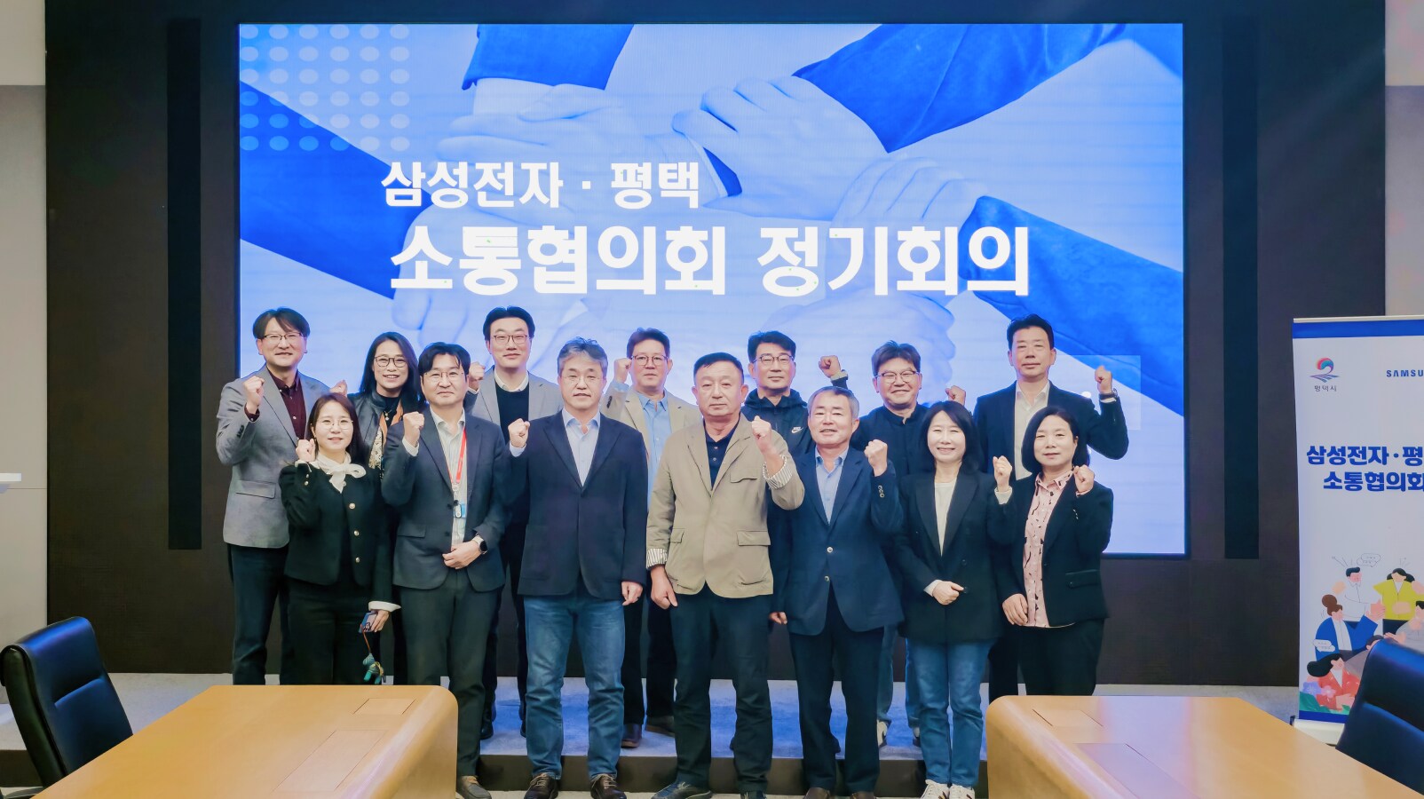 ‘삼성전자·평택 소통협의회 정기회의’ 화면 앞에서 참석자들이 함께 서서 주먹을 쥐고 포즈를 취하고 있다. 모두 밝은 표정으로 단체 사진을 촬영하고 있다.
