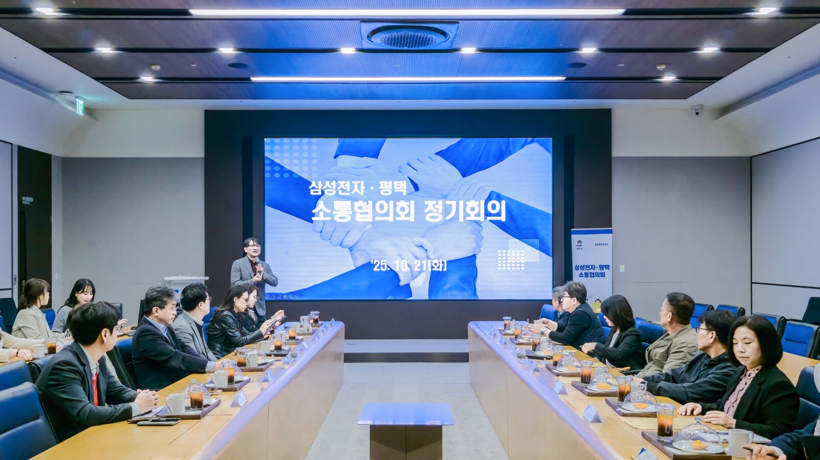 ‘정기 회의에 참석한 소통협의회 위원들의 모습’이라는 문구가 표시된 화면 앞에서 발표자가 마이크를 들고 발언하고 있으며, 양쪽 테이블에 앉은 위원들이 발표를 경청하고 있다.