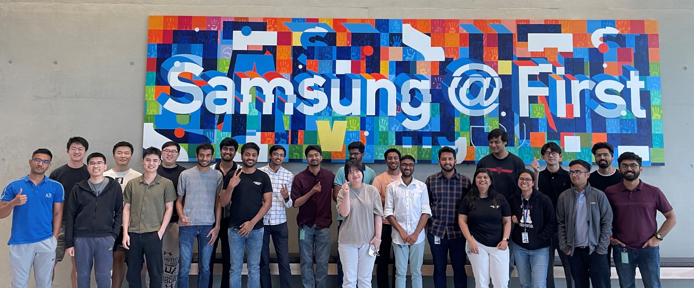 A group of SARC/ACL internship students standing in front of a colorful "Samsung @ First" mural, smiling and posing for a team photo.