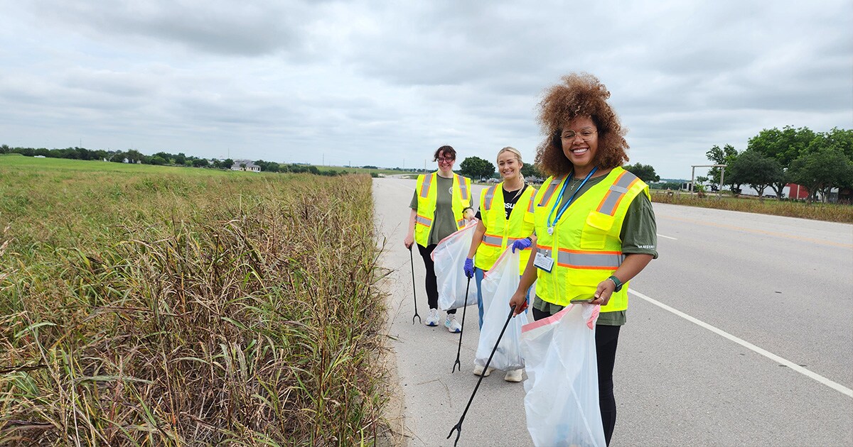 Employees collect litter along a roadside during a volunteer cleanup event supporting local environmental stewardship.