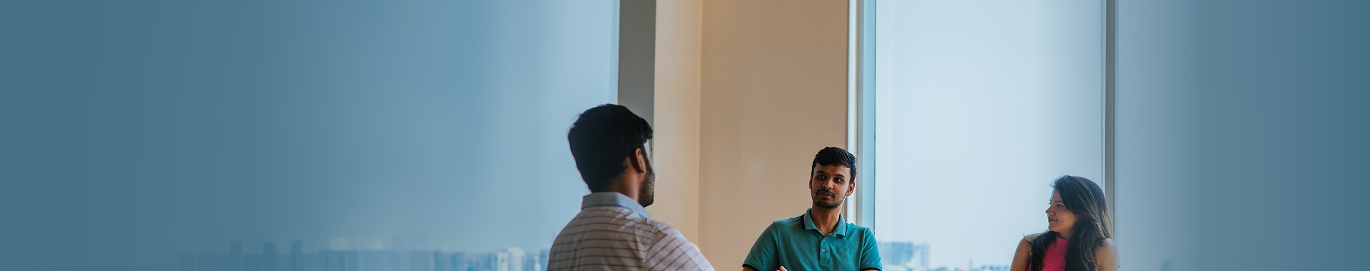 Three professionals engaged in a discussion near a large window with a cityscape view, in a modern office environment.