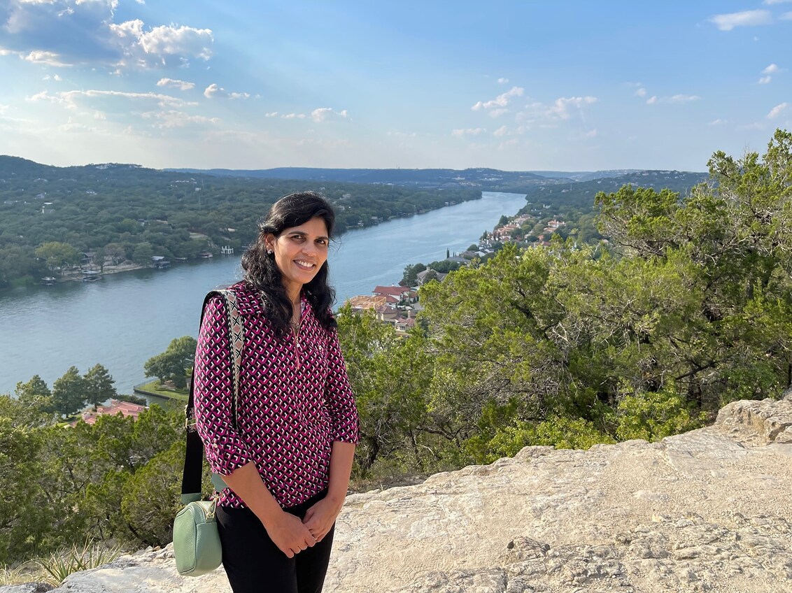 Sudha Nagaraj smiling at a scenic overlook with a river and hills behind her.