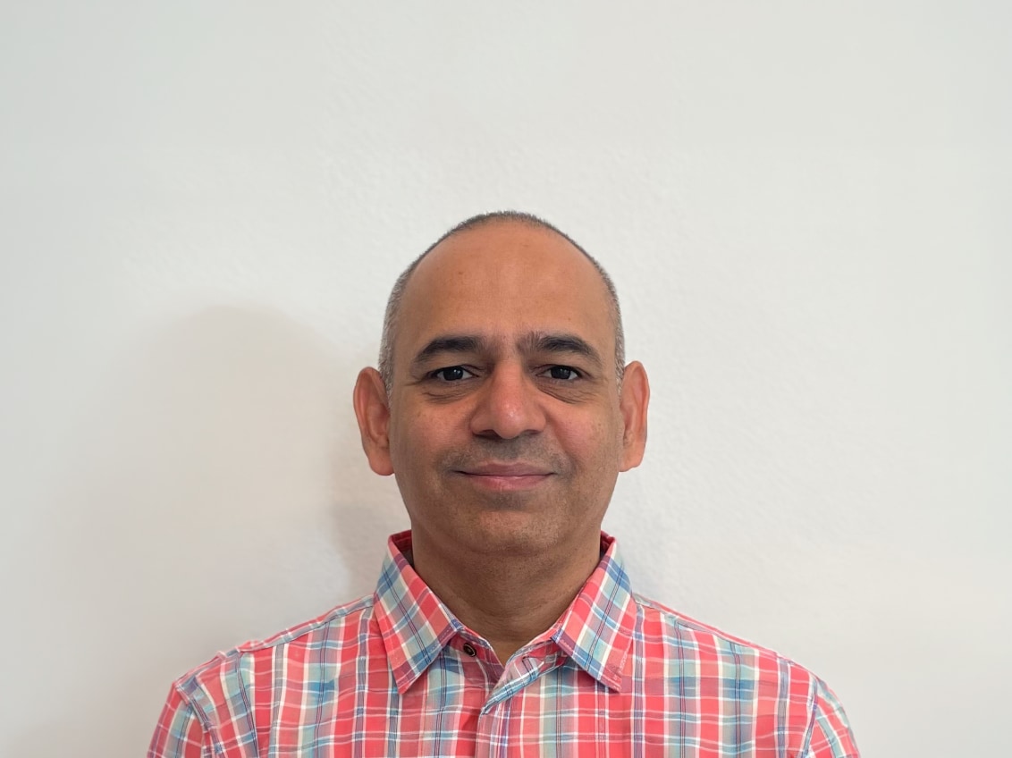 A man with a shaved head wearing a red and blue plaid shirt stands in front of a plain white wall, looking directly at the camera and smiling slightly.