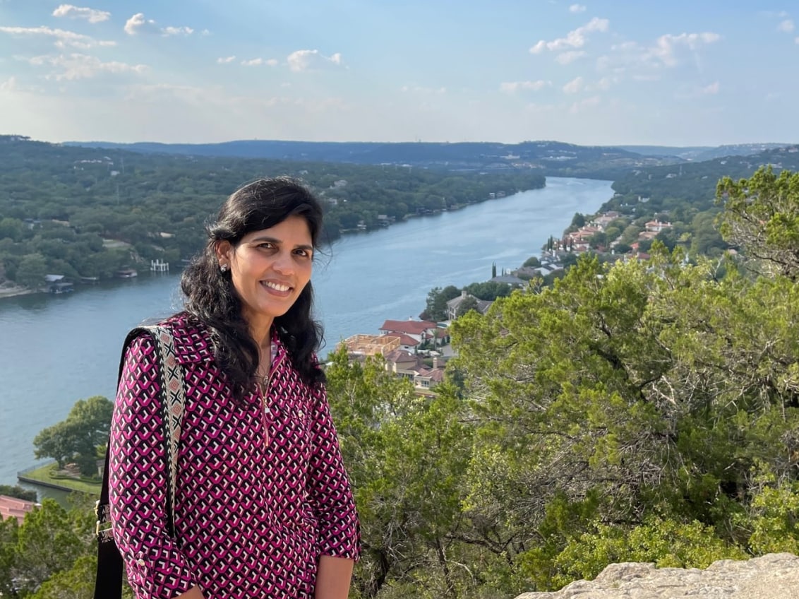 Sudha Nagaraj smiling at a scenic overlook with a river and hills behind her.