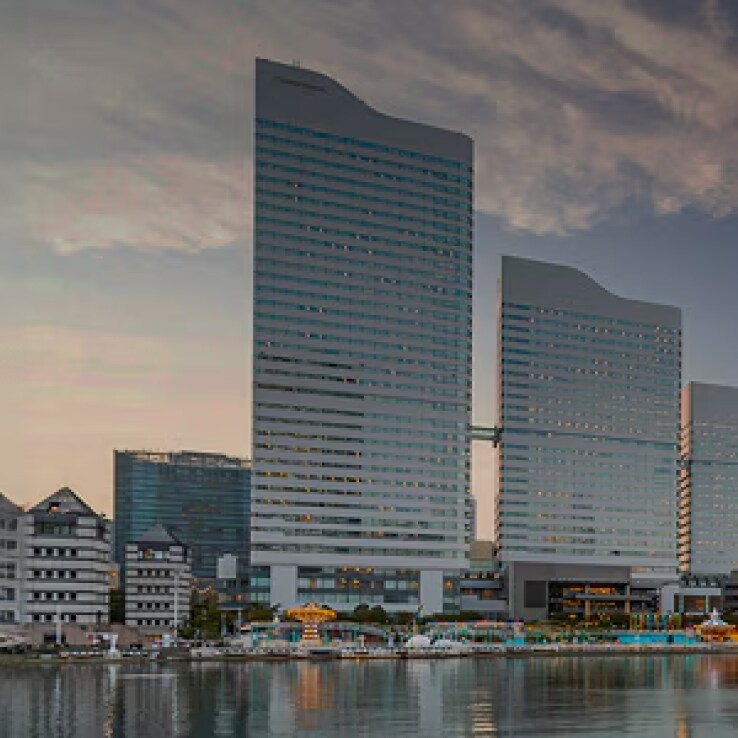Waterfront cityscape at sunset with modern high-rise buildings reflected in the calm water, a large Ferris wheel on the right, and a tree-lined promenade with benches in the foreground.