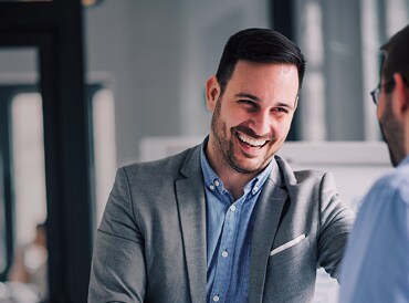 A smiling businessman in a grey blazer greets a colleague in a modern office setting.