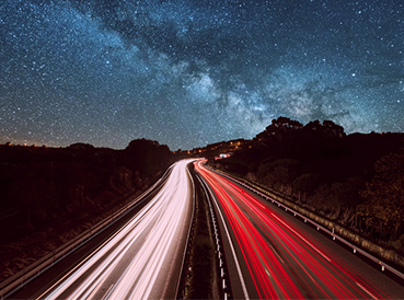 A long-exposure night shot of a highway with red and white light trails under a clear, star-filled sky and the Milky Way.