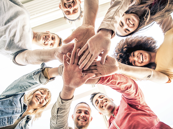 A group of diverse young adults smiling and stacking their hands together in a circle, viewed from below.