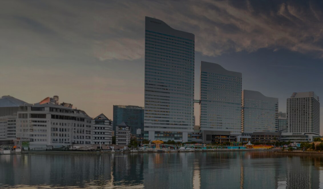Waterfront cityscape at sunset with modern high-rise buildings reflected in the calm water, a large Ferris wheel on the right, and a tree-lined promenade with benches in the foreground.