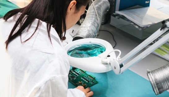 A technician in a white lab coat examining a green circuit board through a magnifying lens in a clean laboratory setting.