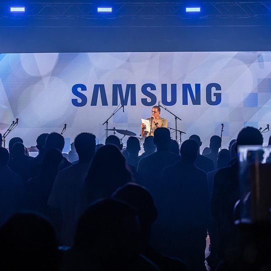Dim event hall filled with a large crowd; an emcee holds a mic in front of a prominent backdrop that reads “Samsung.”