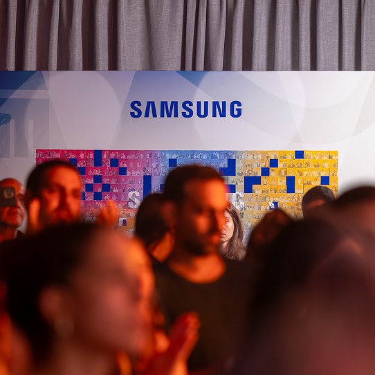 A small, out‑of‑focus group clapping, while a “Samsung” photowall dominates the scene, partially obscured by the people.