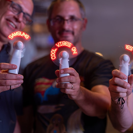Three employees hold an electric fan that displays LED messages “Samsung” and “Cool Vibes Only.”
