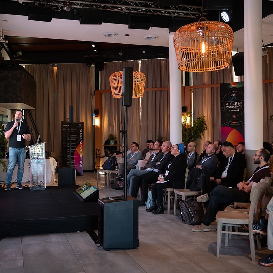 Employees listening to a presentation by a presenter in casual clothing, set in a dimly‑lit hall.