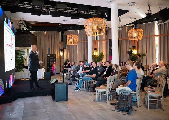 Employees listening to a presentation by a presenter in a suit, set in a dimly‑lit hall