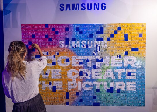 An employee is posting a photo on a large colorful mosaic‑style photowall composed of other employee photos, bearing the phrase “Samsung – Together We Create the Picture.”