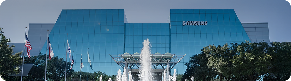 The Samsung Austin campus in Texas, featuring a mirrored glass façade, a central fountain, and national and state flags in the foreground.