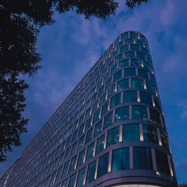 A curved glass office building photographed at dusk, with lights glowing from within and framed by silhouetted tree branches.