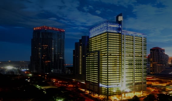 A brightly lit office tower in Southeast Asia, glowing against the night sky with surrounding skyscrapers and city lights.