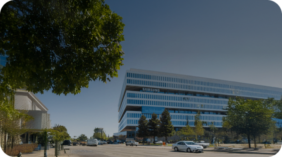 The Samsung corporate building in the U.S., captured on a sunny day with clear skies, cars parked nearby, and leafy trees in the foreground.